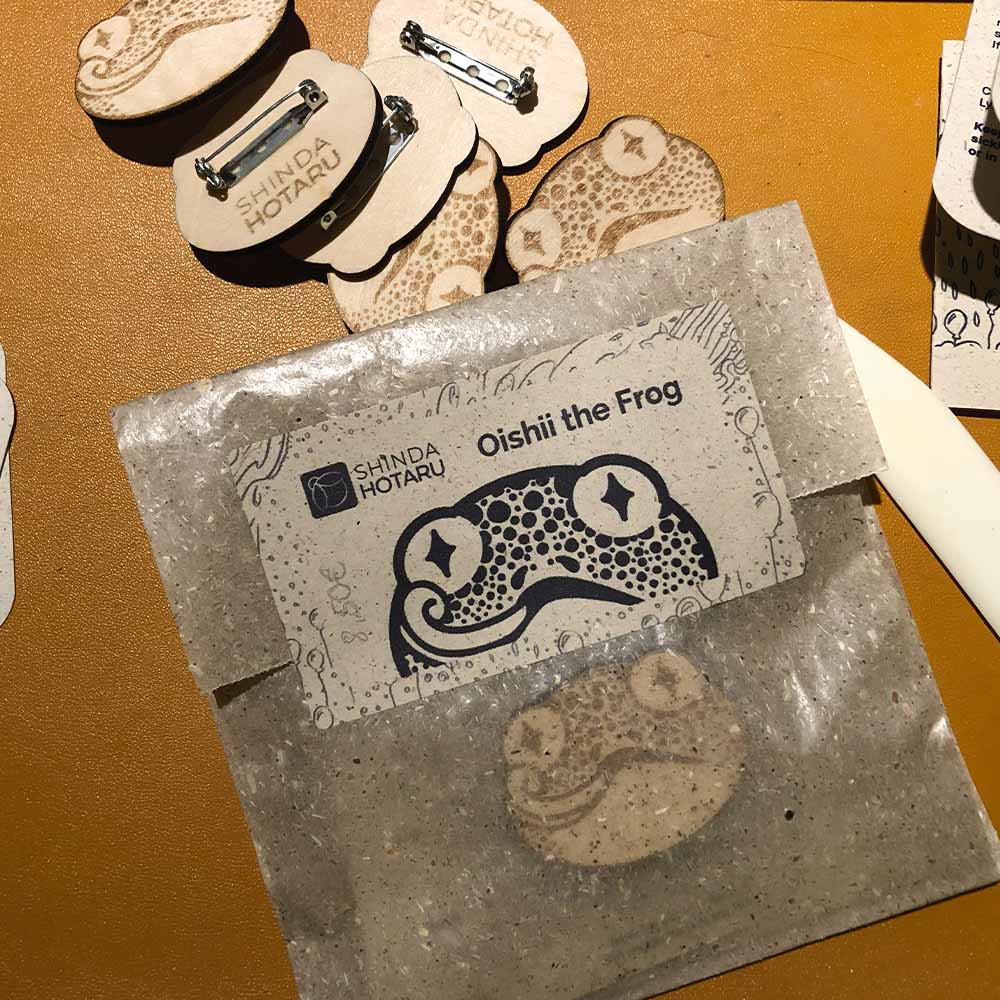 Ein in Pergamin-Tüte verpackter Holzanstecker. Als Verschluss dient ein Aufkleber, auf dem das Motiv ist.
