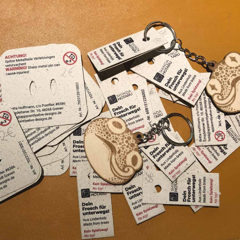 Verschiedene Etiketten und Backing Cards. Zwei Holzs-Schlüsselanhänger mit jeweils einem Froschmotiv liegen auf den dazugehörigen Etiketten.